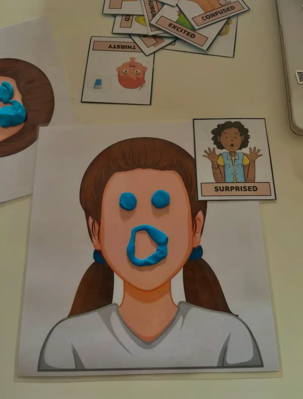 Cara de plastilina con expresión de sorpresa junto a tarjetas de emociones en inglés